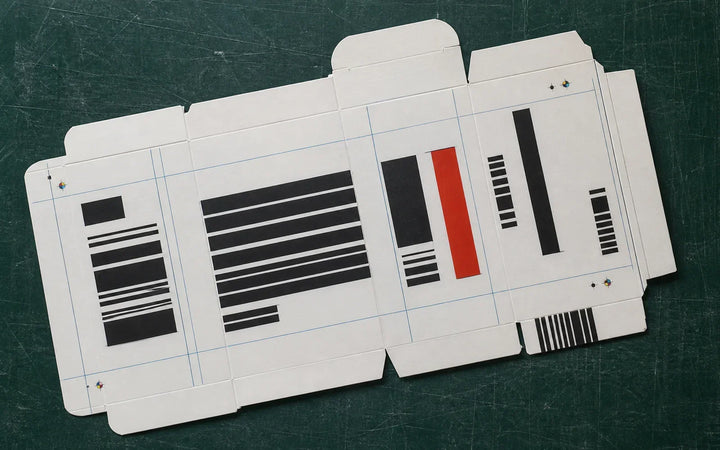 Unfolded white carton proof with black bars and a red panel on a green cutting mat